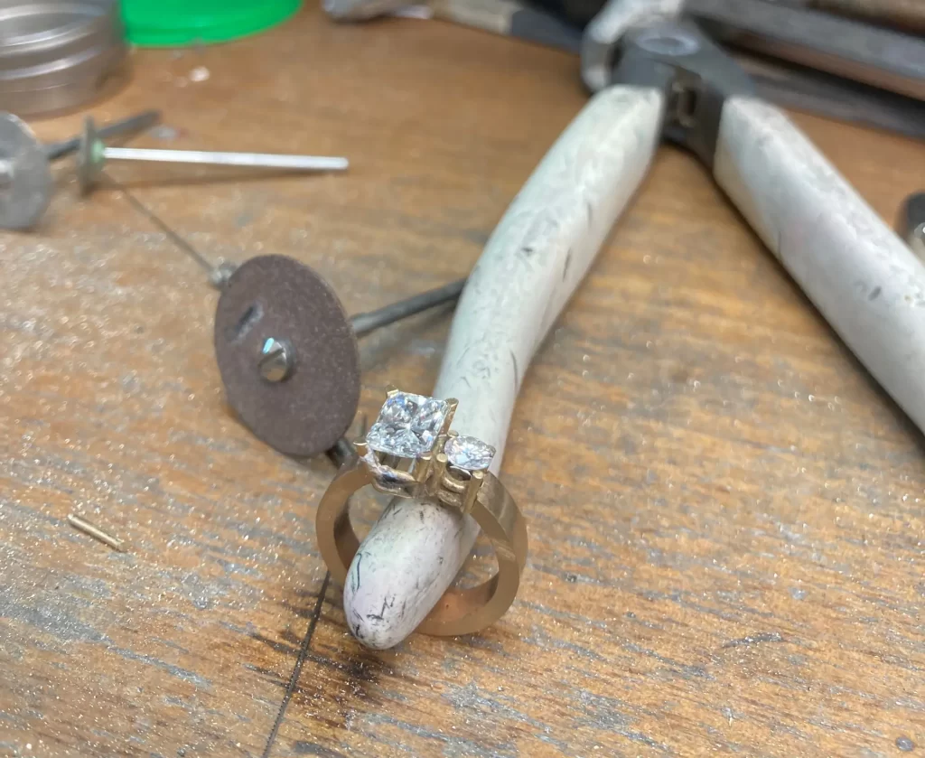 Our Process 4 A diamond ring placed on a pair of pliers in a jeweler’s workshop, surrounded by various jewelry tools and equipment on a wooden workbench.