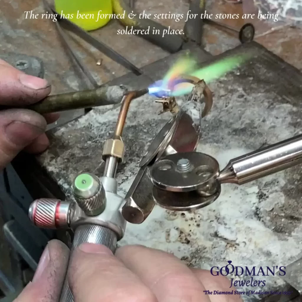 Our Process 3 A jeweler carefully soldering the settings for gemstones onto a ring using a torch in a workshop, with tools and equipment in the background.